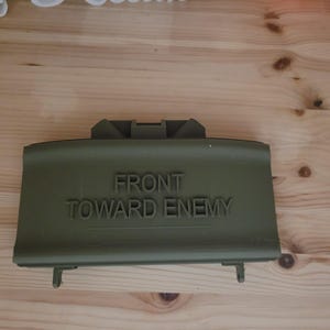 May include: An olive green, rectangular object with the words "FRONT TOWARD ENEMY" embossed on the top. The object has a raised handle and two small protrusions on the bottom. The background is a light-coloured wooden surface.