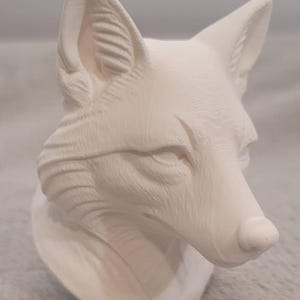 May include: A matte white 3D printed fox head bust featuring visible horizontal layer lines across the surface. The sculpture displays detailed fur texture, pointed ears, and a defined snout, capturing the fox in a forward-facing profile.