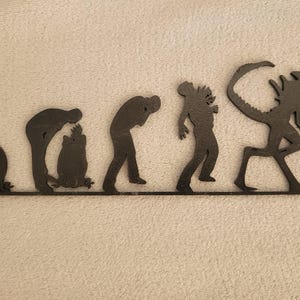 May include: A black wall decoration featuring a five-stage evolutionary sequence. It begins with an egg, followed by a facehugger, a chestburster, a human figure, and finally a full-grown xenomorph creature, all connected by a single base line.