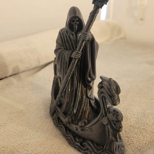 May include: A black 3D-printed figurine of a hooded skeletal figure standing in a boat. The figure holds a long staff topped with a skull. The boat features a dragon-shaped prow and is decorated with small skulls along the sides and wavy water details.
