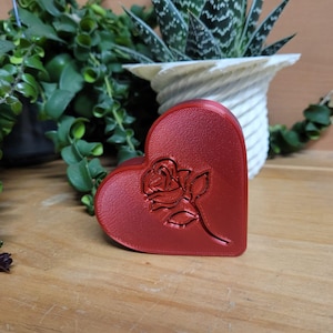 May include: A red heart-shaped leather item with a rose design. The embossed rose is centered on the front of the heart. The item is on a wooden surface, with green plants and a white pot in the background.