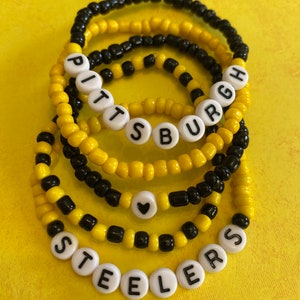 May include: A set of six black and yellow beaded bracelets with white beads spelling out "PITTSBURGH STEELERS" and a small black heart charm.