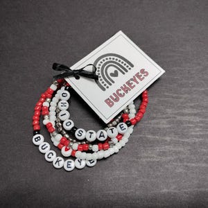 May include: A set of five beaded bracelets with the words "Ohio State Buckeyes" spelled out in white beads with red and black accents. The bracelets are on a white card with a rainbow and the word "Buckeyes" printed on it.