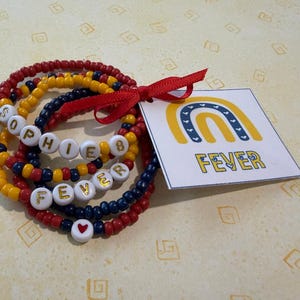 May include: A stack of beaded bracelets in red, yellow, and navy blue, featuring white letter beads spelling SOPHIE B and FEVER, a small red heart bead, and a red ribbon tied to a square tag with a yellow and blue rainbow graphic and the text FEVER.