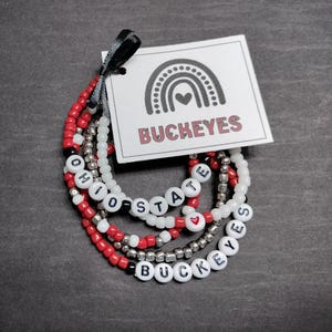 May include: Set of four beaded bracelets with the words "Ohio State" and "Buckeyes" in black lettering on white beads. The bracelets are red, white, and silver.