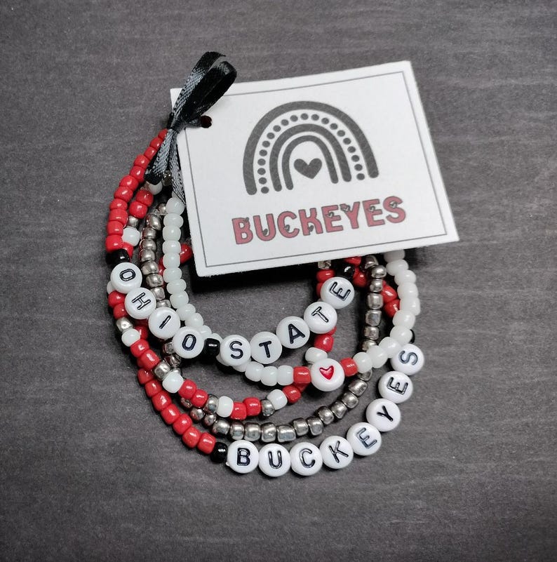 May include: Set of four beaded bracelets with the words "Ohio State" and "Buckeyes" in black lettering on white beads. The bracelets are red, white, and silver.