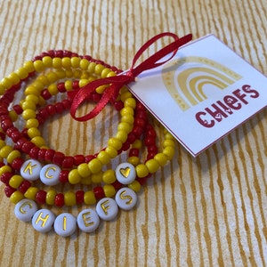 May include: A set of six beaded bracelets in red and yellow with white beads spelling out "CHIEFS" and a heart charm. The bracelets are tied together with a red ribbon and a card with a rainbow design and the word "CHIEFS" in red.