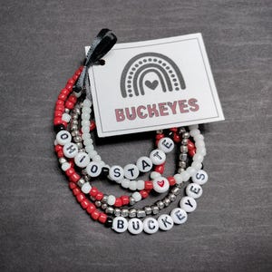 May include: Set of five beaded bracelets with the words "Ohio State" and "Buckeyes" spelled out in black letters on white beads. The bracelets are red, white, and silver.