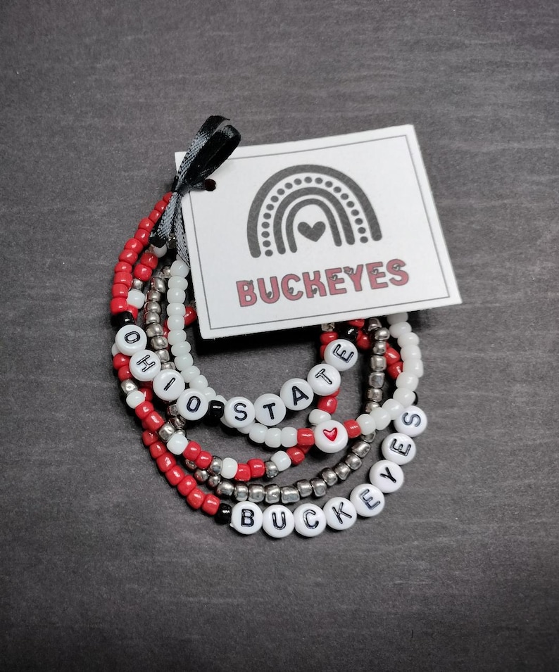 May include: Set of five beaded bracelets with the words "Ohio State" and "Buckeyes" spelled out in black letters on white beads. The bracelets are red, white, and silver.