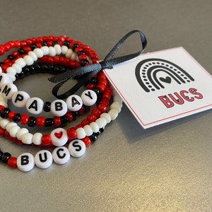 May include: Stack of beaded bracelets in red, white, and black. One bracelet spells out "TAMPA BAY" and another "BUCS" with a heart. A tag with a rainbow and the word "BUCS" is attached with a black ribbon.