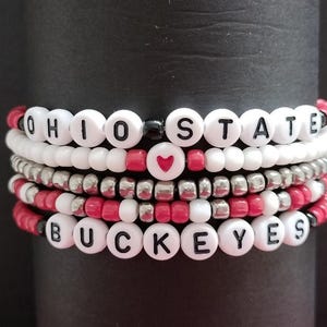 May include: A stack of beaded bracelets with the words "Ohio State" and "Buckeyes" spelled out in black letters on white beads. The bracelets are interspersed with red and silver beads. A single red heart bead is visible in the center of the stack.