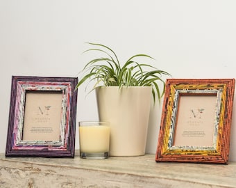 May include: Two decorative picture frames made of rolled paper with colorful patterns. The frames are on a wooden surface with a potted plant and a candle in front of them. The frames have a small card inside with the text "Memento Home".