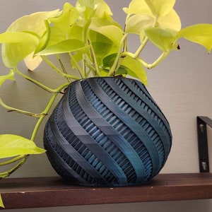 May include: A bright green plant with heart-shaped leaves in a dark blue, textured, spherical pot. The pot is on a dark brown wooden shelf, supported by black metal brackets, against a grey wall.