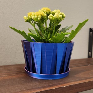 May include: A small potted plant with yellow flowers in a blue and purple iridescent planter. The planter has a geometric design and is sitting on a wooden shelf.