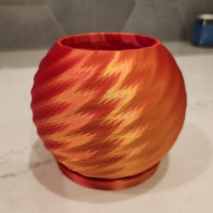 May include: A red and orange 3D printed planter with a swirling pattern. The planter has a round base and a wide, open top.