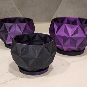 May include: Three geometric planters in different sizes, made of a dark purple and black plastic. The planters have a geometric design and are perfect for adding a modern touch to any home.