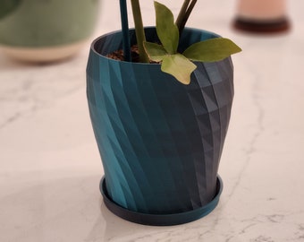 May include: A teal blue planter with a geometric pattern. The planter has a black plastic saucer. A small green plant is growing inside the planter.