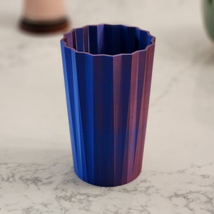 May include: A tall, cylindrical container with a fluted design. The colour gradient transitions from a deep blue to a vibrant purple. The container has a ridged, zig-zag top edge and is likely made of plastic or a similar material.