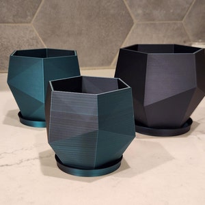 May include: Three geometric planters in varying sizes and shades of teal and black. Each planter has a faceted design and sits on a matching round saucer. The planters are made of a textured material.
