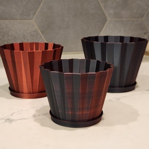 May include: Three decorative plant pots in varying sizes and shades of brown and black. Each pot has a ridged, geometric design and sits on a matching saucer. The pots are arranged on a white countertop.