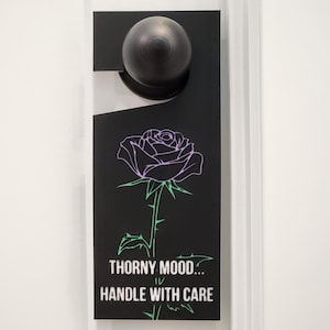 May include: Black door hanger with a rose illustration in purple and green. The text reads "Thorny Mood... Handle With Care". The door hanger is hanging on a white door with a dark metal doorknob.