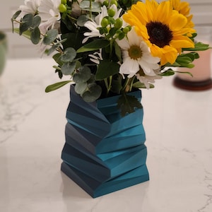 May include: A blue geometric vase with a white and yellow flower arrangement. The vase is made of a textured material and has a modern design.