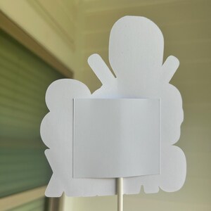 Op de afbeelding: Witte papieren uitsnijding van een cartoonfiguur met een leeg wit vierkant in het midden. De uitsnijding is bevestigd aan een witte stok.