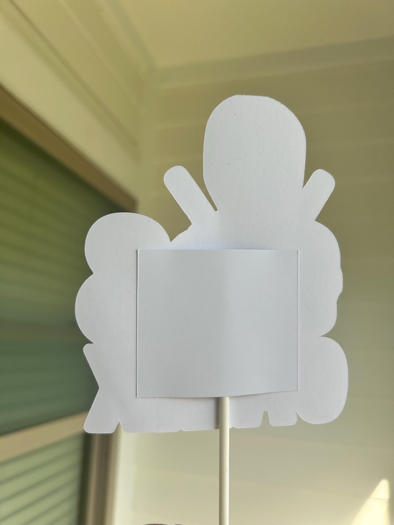 Op de afbeelding: Witte papieren uitsnijding van een cartoonfiguur met een leeg wit vierkant in het midden. De uitsnijding is bevestigd aan een witte stok.