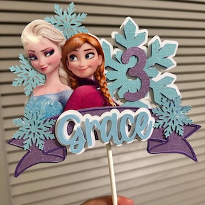 May include: A cake topper featuring the characters Elsa and Anna from the movie Frozen. The topper is decorated with snowflakes and a purple banner with the name "Grace" and the number "3".