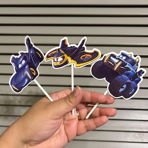 May include: Three blue and black cartoon vehicle cupcake toppers. The toppers feature a monster truck, a jet, and a car. The toppers are on white sticks.