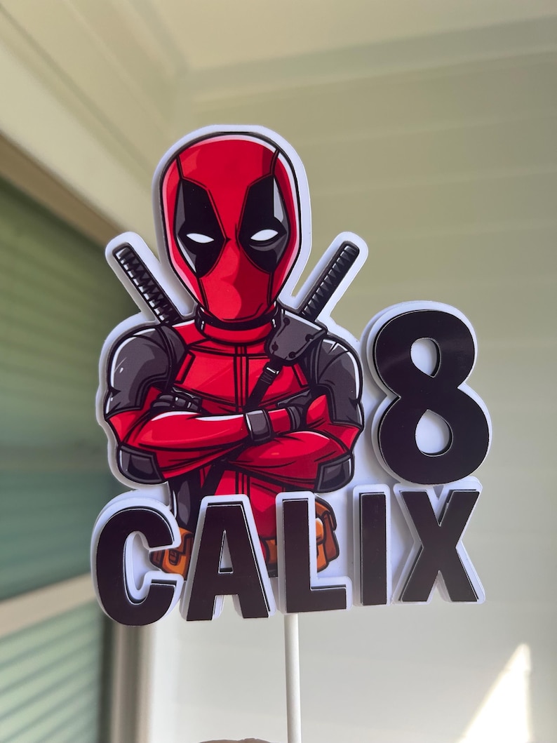 Op de afbeelding: Een rode en zwarte Deadpool-taarttopper met het nummer 8 en de naam Calix. De topper is gemaakt van karton en is perfect voor een verjaardagstaart.