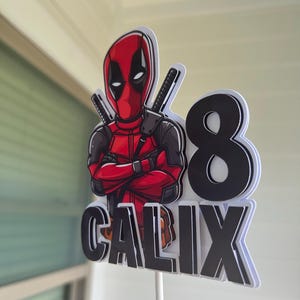 Op de afbeelding: Een rode en zwarte Deadpool-taarttopper op een witte achtergrond. De topper toont Deadpool met gekruiste armen, een zwart-wit zwaard en het nummer 8. De naam "CALIX" staat in zwarte letters onder het nummer.