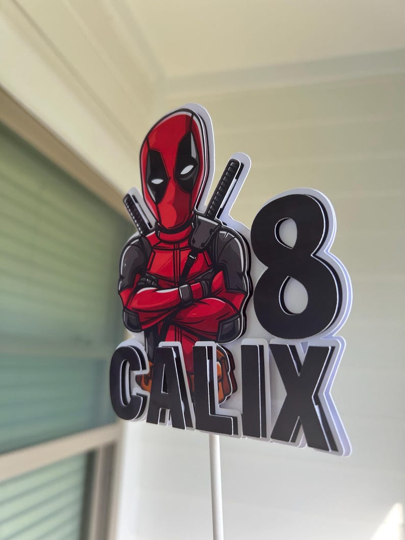 Op de afbeelding: Een rode en zwarte Deadpool-taarttopper op een witte achtergrond. De topper toont Deadpool met gekruiste armen, een zwart-wit zwaard en het nummer 8. De naam "CALIX" staat in zwarte letters onder het nummer.