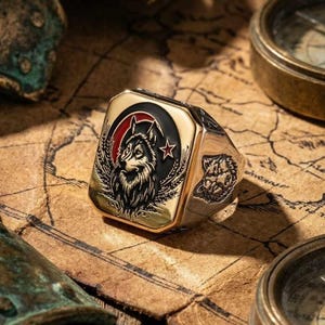 Puede incluir: Un anillo de oro y plata con un diseño de lobo. El anillo presenta una cara rectangular con un retrato detallado de un lobo, una media luna roja y una estrella. El anillo está sobre un mapa.