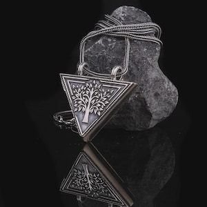 May include: A triangular silver pendant featuring an embossed tree design with intricate branches and leaves. The pendant hangs from a woven silver chain, showcasing a polished metallic finish with darkened recessed details for contrast.