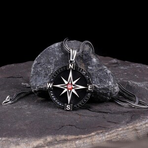 May include: A silver compass pendant necklace with a black background. The compass rose features a red gemstone center and is surrounded by black and clear stones. The necklace chain is also silver.