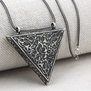 Puede incluir: Un collar de plata con un colgante triangular con un intrincado diseño floral. El colgante está unido a una cadena de plata.
