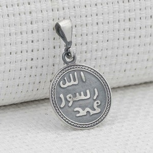 May include: Silver pendant with a round, grey, engraved charm featuring Arabic script. The charm is surrounded by a silver border with a decorative pattern.