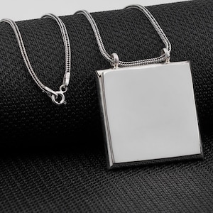 May include: A silver necklace with a square pendant. The pendant is a polished, reflective square. The chain is a woven design. The necklace is displayed on a black textured surface.