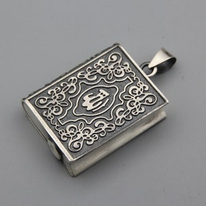Puede incluir: Colgante de plata con forma de libro pequeño. El colgante tiene intrincados diseños florales y de volutas en la cubierta y una anilla para sujetarlo a una cadena. La cubierta del libro presenta un diseño central.