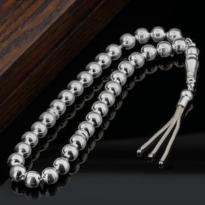 May include: A string of polished, spherical silver-coloured beads arranged in a continuous loop. The beads are uniform in size and reflective. At one end, a decorative silver-coloured metal component connects to a short, three-strand silver tassel.