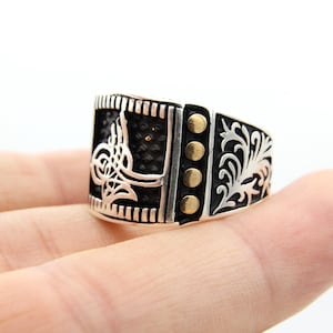 Könnte beinhalten: Silberring mit schwarzem Mittelteil und goldenen Akzenten. Der Ring hat ein verziertes Design an den Seiten und ein dekoratives Emblem auf der Vorderseite. Der Ring wird in einer Hand vor weißem Hintergrund gehalten. Schmuck.