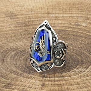 Può includere: Un anello d'argento con un'ampia incastonatura di pietra blu a forma di scudo. La pietra presenta una calligrafia color oro. L'anello ha un design ornato con dettagli a mezzaluna sul lato.