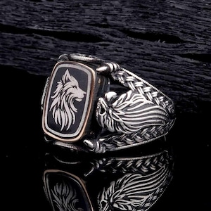 Op de afbeelding: Een zilveren ring met een rechthoekige zwarte voorkant met een wolfontwerp. De ring heeft een gedetailleerde gevlochten band en een goudkleurige rand rond de wolfafbeelding. De ring wordt op een donkere ondergrond getoond.