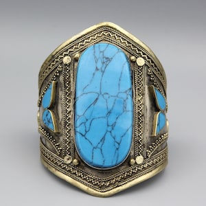 May include: A wide, ornate cuff bracelet with a large, oval turquoise stone at the center. The bracelet has a gold-colored metal frame with intricate detailing and smaller turquoise accents. The design is symmetrical and features a vintage or ethnic aesthetic.