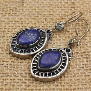 May include: A pair of silver-toned earrings with oval, dark blue lapis lazuli stones. The earrings have a decorative border and a hook closure. The background is a textured, tan fabric.