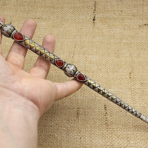 Może przedstawiać: Ozdobna srebrna różdżka ze złotymi i czerwonymi akcentami. Różdżka ma dekoracyjny uchwyt z motywem w kształcie serca i jest ozdobiona trójkątnymi i diamentowymi wzorami. Różdżka jest trzymana w dłoni.