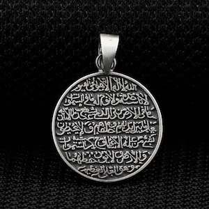 May include: A circular, silver-toned pendant with a loop for a chain. The pendant is inscribed with detailed Arabic script. The background is a dark, textured material.