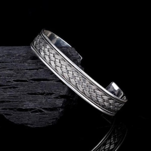 May include: A silver cuff bracelet with a woven design. The bracelet is made of a shiny metal and features a detailed pattern across the top. The cuff is resting on a dark, textured surface against a black background.