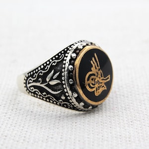 May include: A silver ring with an oval black onyx stone featuring a gold-coloured ornate design. The ring has detailed silver filigree and a gold-coloured border around the black stone.